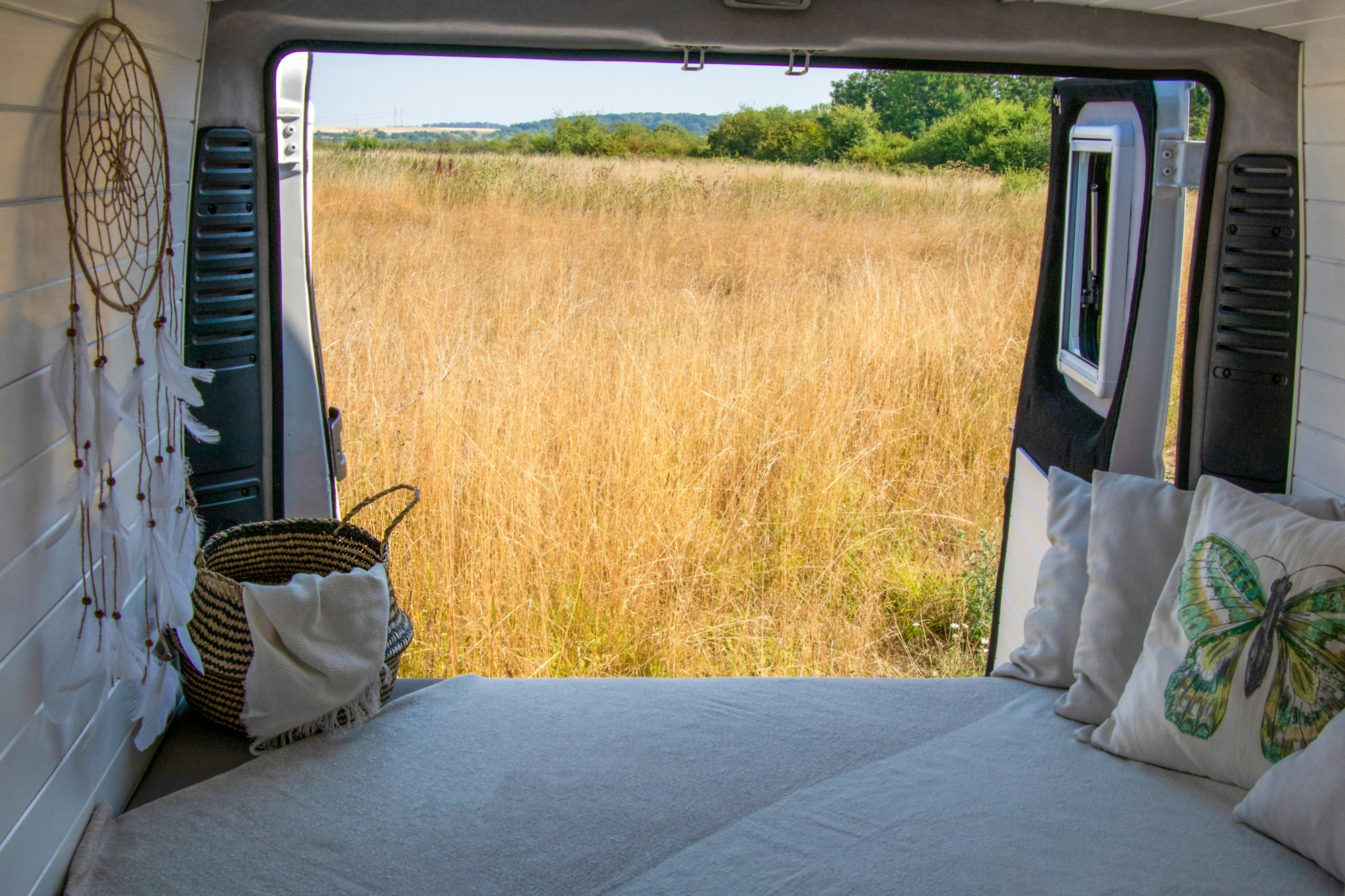 Offene Hecktüren eines ausgebauten Campers mit Matratze, dahinter ein Feld im Abendlicht